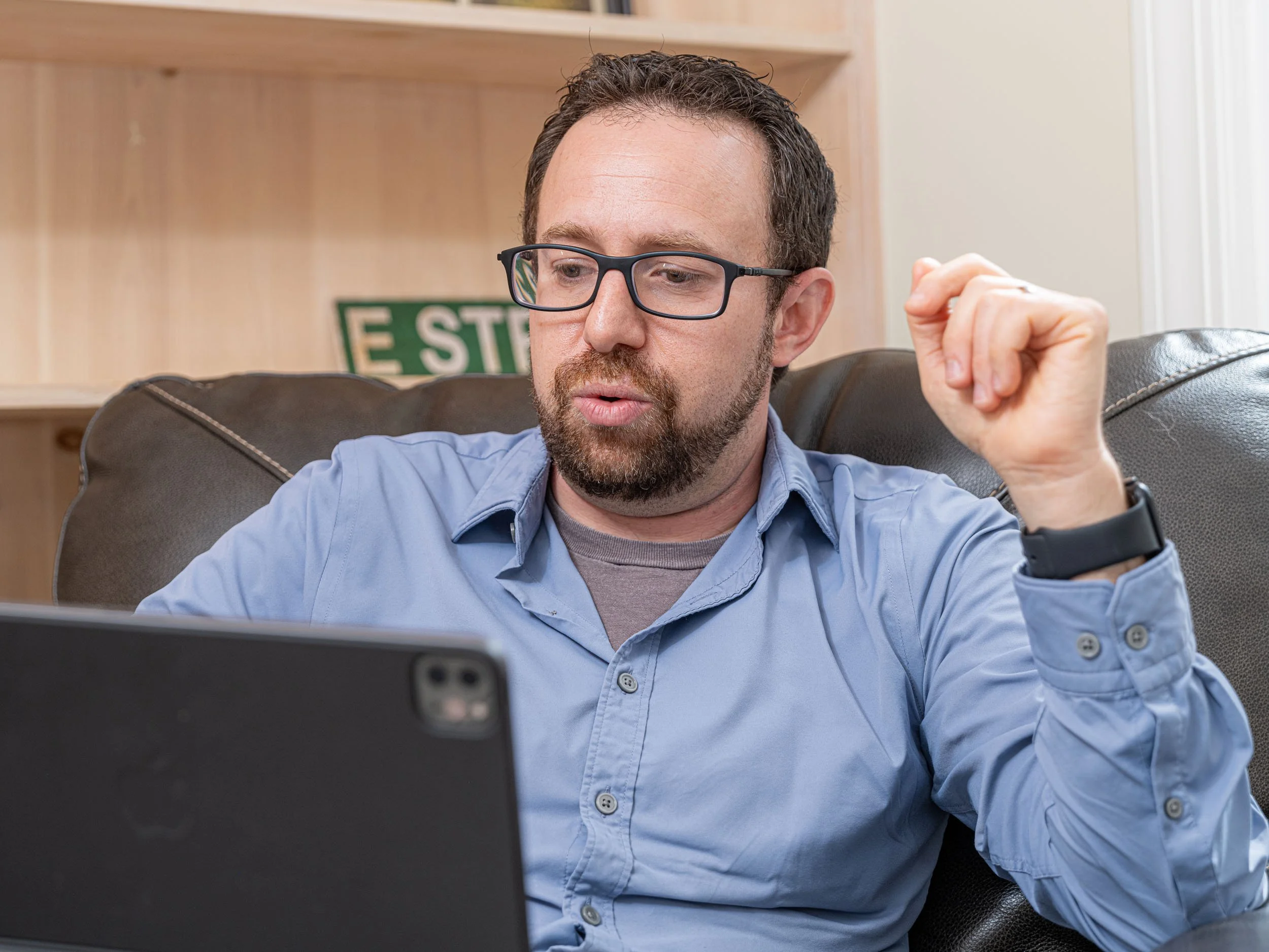Man with glasses and beard sitting on a sofa, looking at a laptop, with a hand raised near his head in a gesture of explanation or emphasis.