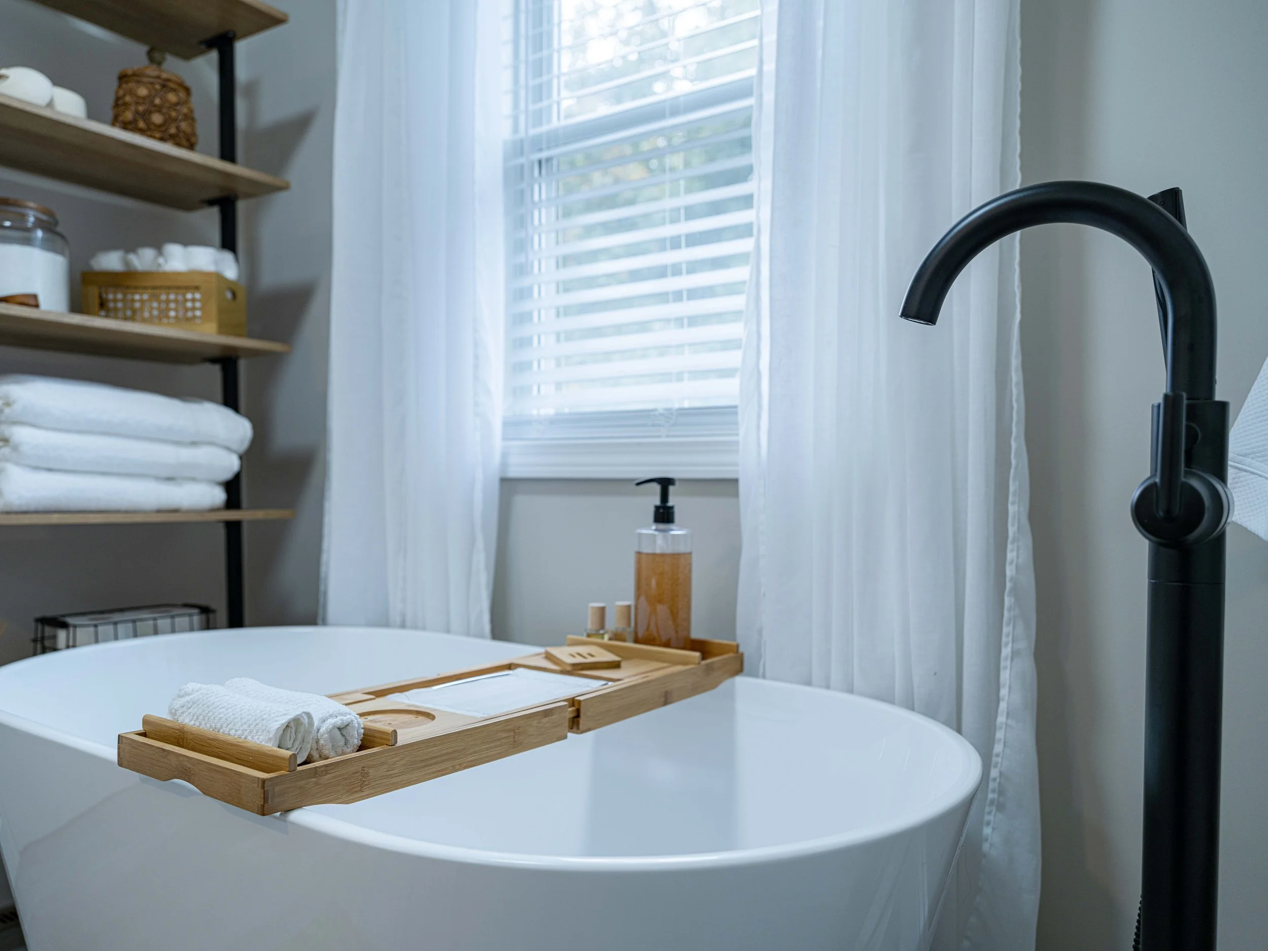 Modern bathroom with a white bathtub, black faucet, wooden bath tray holding rolled white towel and toiletries, white curtains, and shelves with towels and decor.