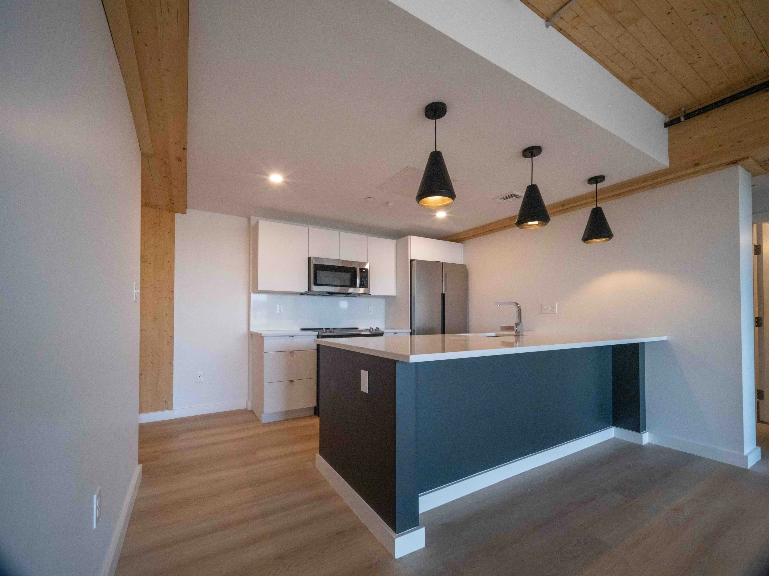 Modern kitchen with white cabinetry, stainless steel refrigerator and microwave, black pendant lights over a white countertop island, wooden ceiling accents, and hardwood floors.