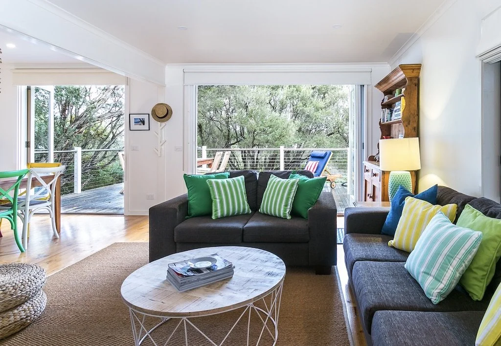 Living room with dark gray sofas, colorful striped and solid pillows, wooden coffee table, sliding glass door opening to a balcony with outdoor seating, and trees in the background.