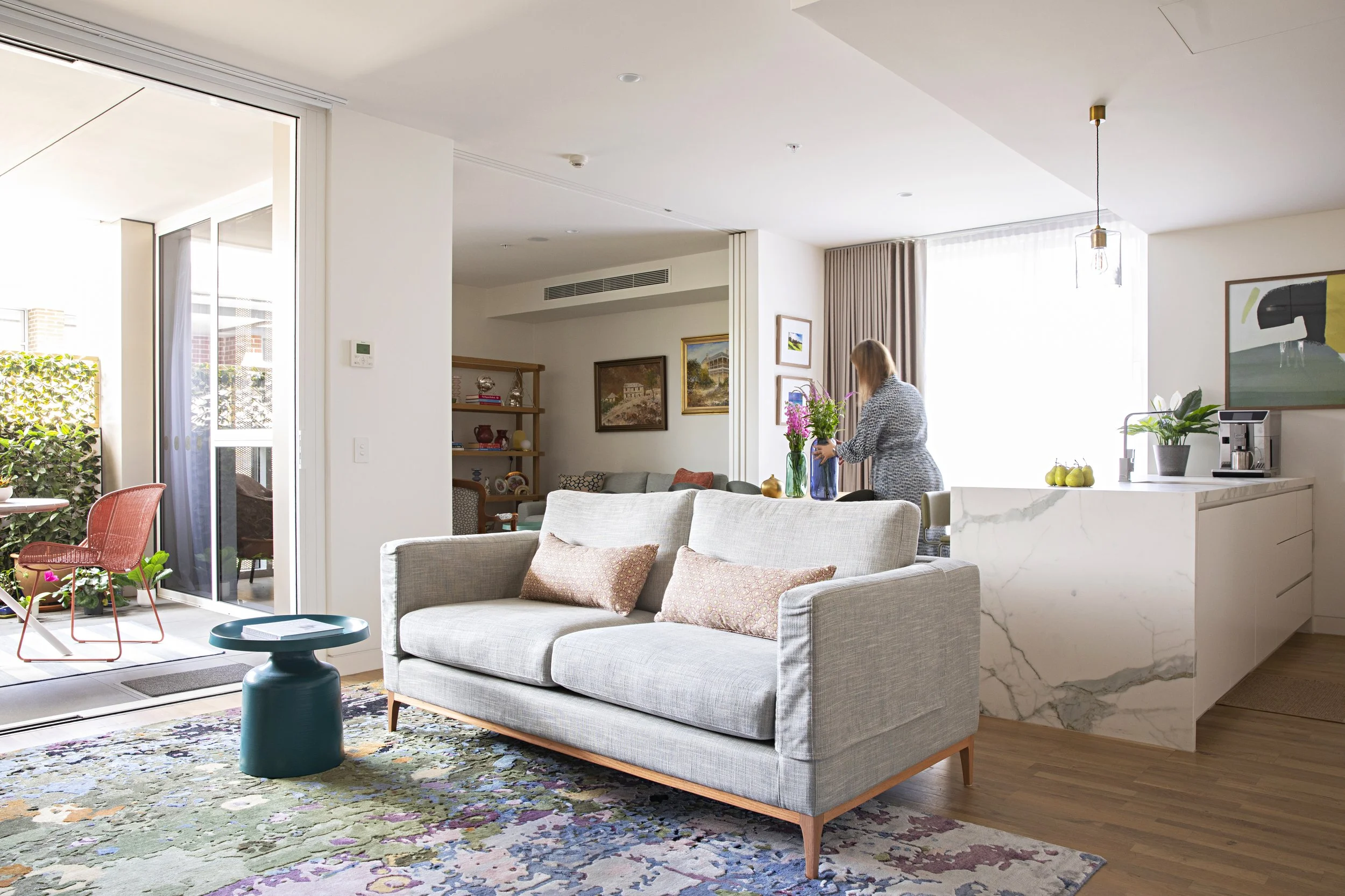 Modern living room with a beige sofa, colorful abstract rug, and large sliding glass doors opening to a balcony with outdoor seating and greenery. A woman arranges flowers on the kitchen counter with artwork on the walls, and natural light fills the space.
