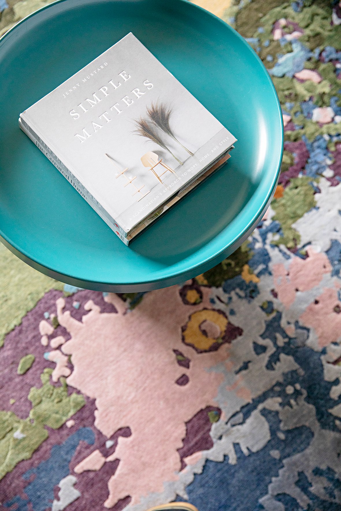 A hardcover book titled 'Simple Matters' by Jenny Mustard resting on a teal round table, with a colorful abstract patterned rug underneath.
