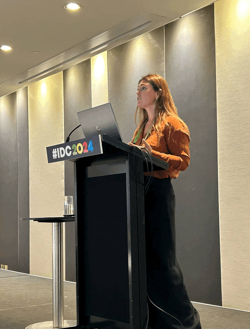A woman giving a presentation at a conference, standing behind a black podium with a laptop. The podium has a sign that reads '#IDC2024'. The room has neutral-colored walls and ceiling lights, with a glass of water on a small side table beside the podium.