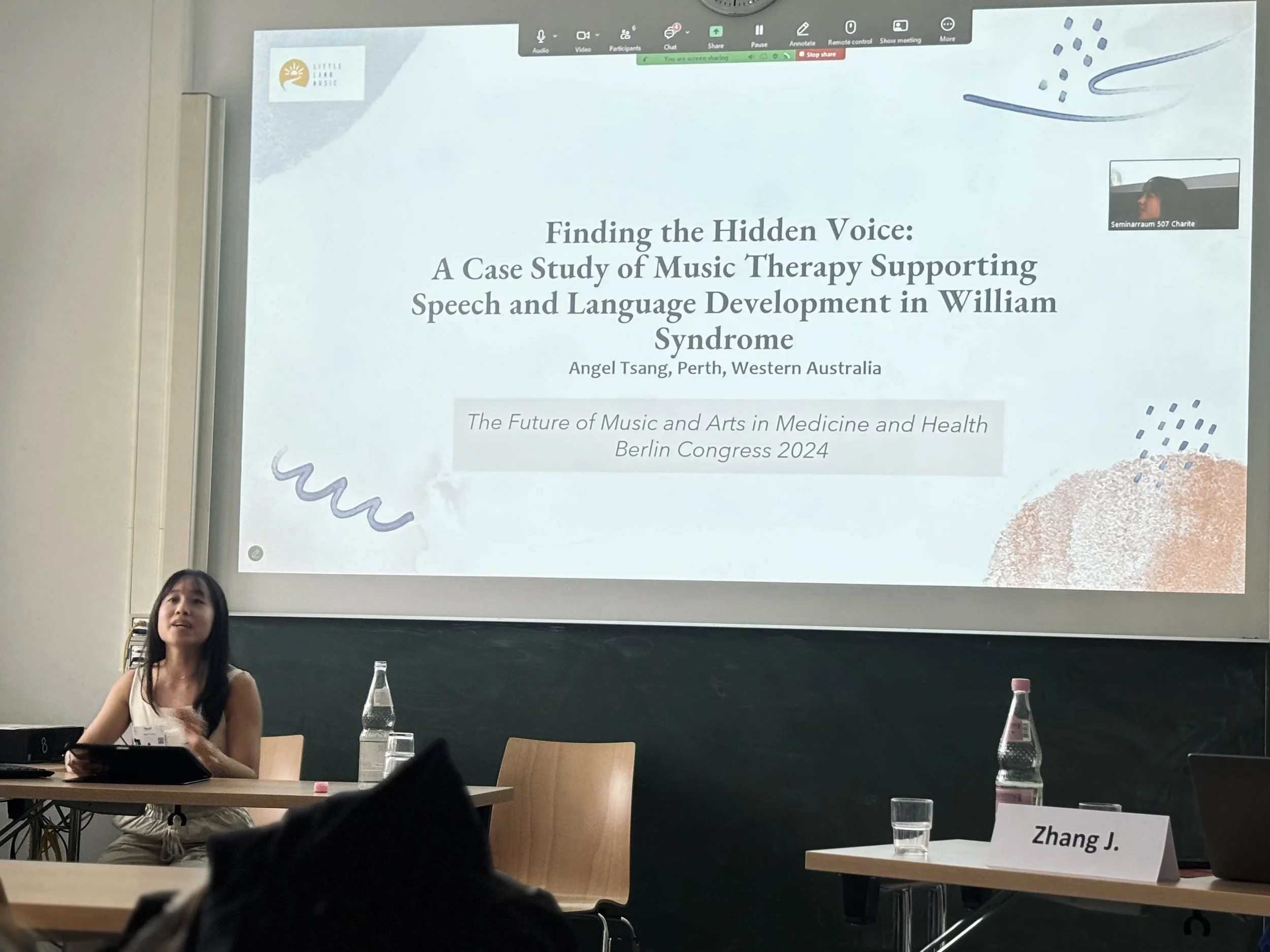 A woman sitting at a desk in a classroom or conference room, speaking into a microphone with water bottles and papers in front of her. A large presentation slide is projected on the wall behind her, displaying the title of a research study about music therapy and William syndrome, with the name Angel Tsang and details about a Berlin congress 2024. The slide includes logos and decorative elements.