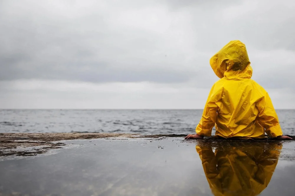 A child in a yellow raincoat sitting by the sea
