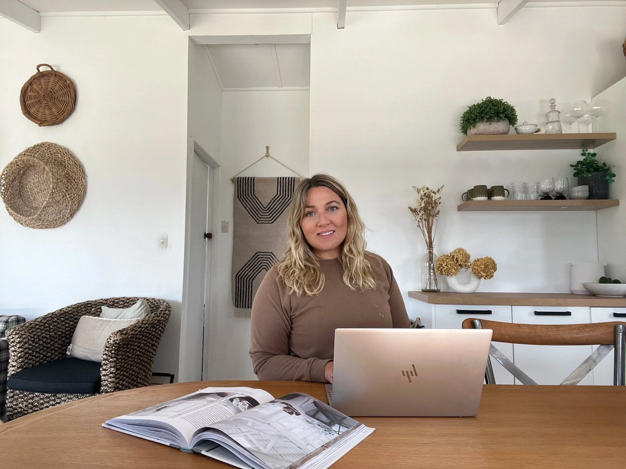 A woman working on a laptop at a wooden table in a cozy, well-decorated room with shelves holding plants, glassware, and decorative items. An open magazine is on the table, and wicker chairs are nearby.