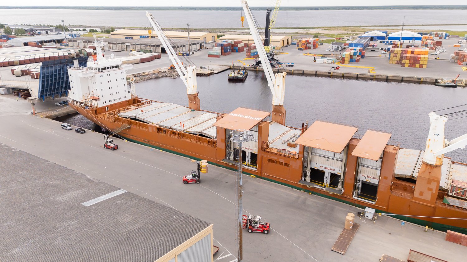 Aerial footage of forklifts, equipped with paper roll clamps, precisely handle and transport large cardboard reels to ship