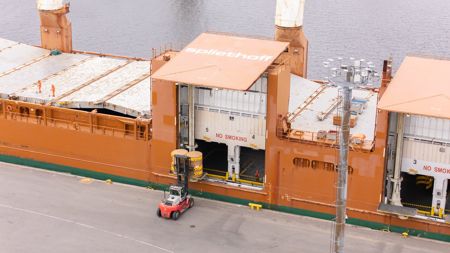 Aerial footage of forklifts, equipped with paper roll clamps, precisely handle and transport large cardboard reels to ship