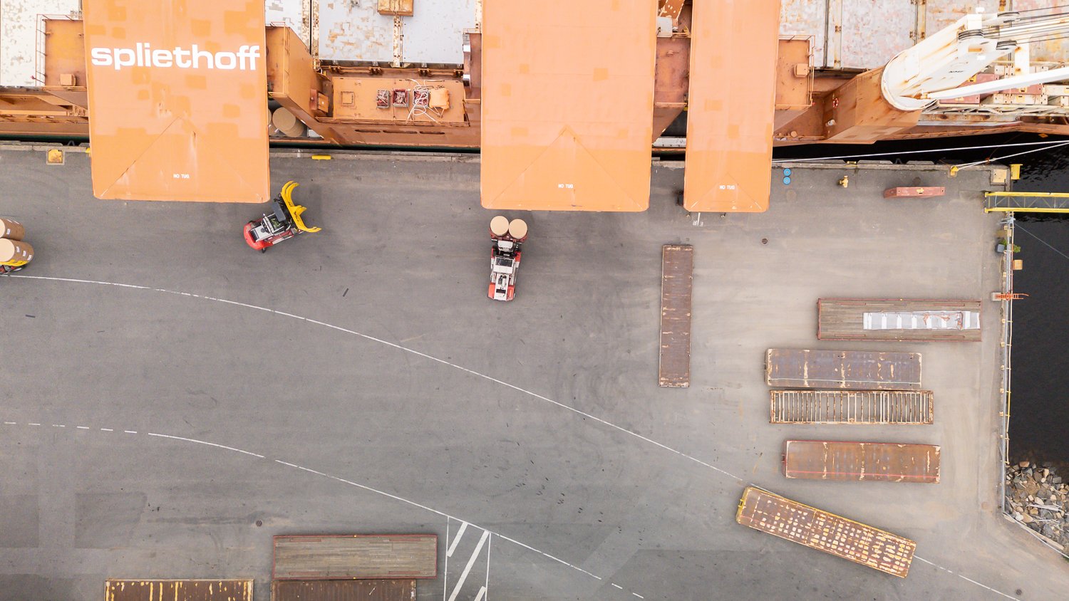 Aerial footage of forklifts, equipped with paper roll clamps, precisely handle and transport large cardboard reels to ship