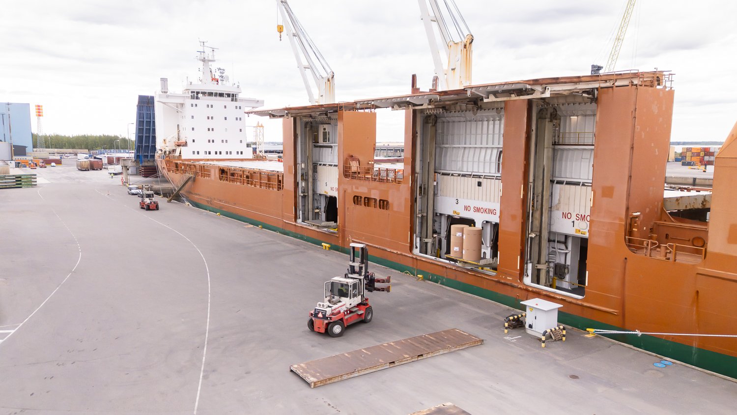Aerial footage of forklifts, equipped with paper roll clamps, precisely handle and transport large cardboard reels to ship