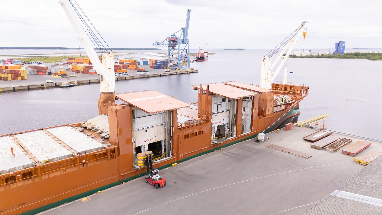 Aerial footage of forklifts, equipped with paper roll clamps, precisely handle and transport large cardboard reels to ship