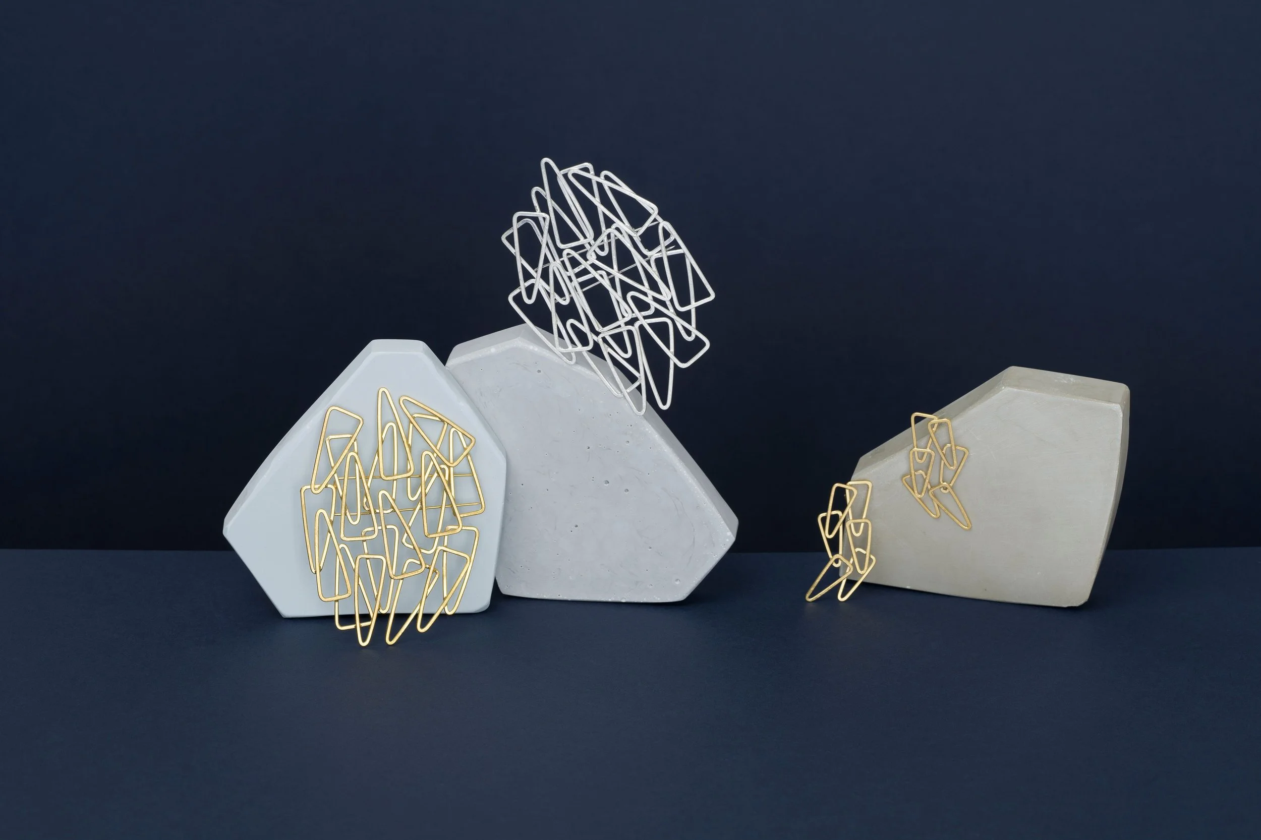 Decorative art pieces with geometric shapes, including white and beige pentagonal and hexagonal bases, adorned with metallic paperclip-like objects in gold and silver, arranged on a dark background.