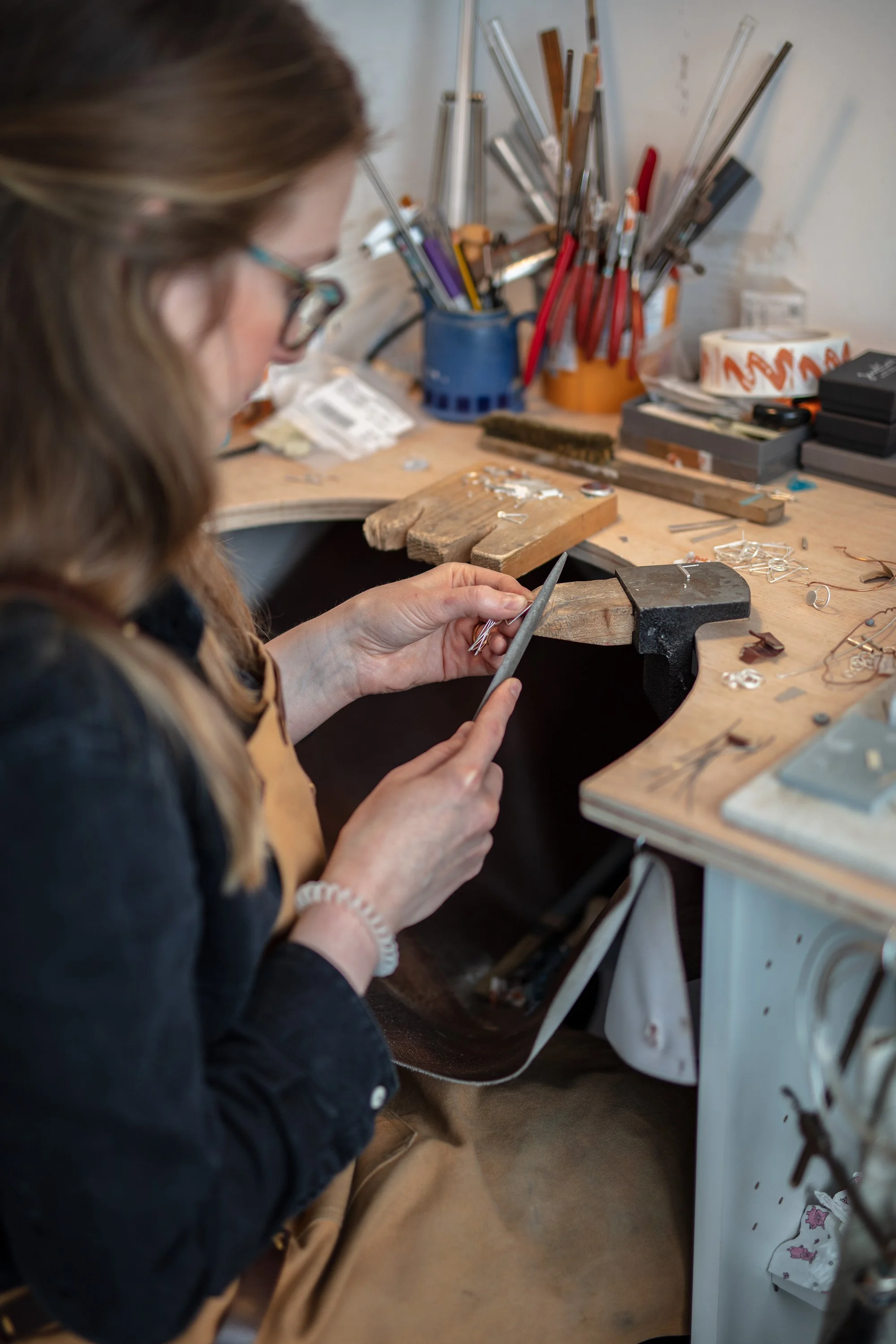 A woman working at a cluttered workbench with jewelry-making tools, metal rings, and various supplies.