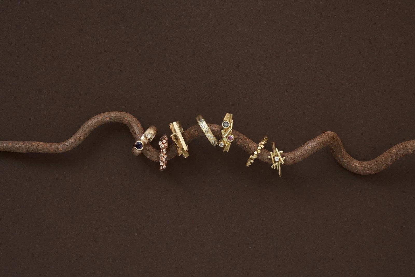 A twisting, brown, textured branch with multiple gold and silver rings with various gemstones and designs placed on it, against a dark background.