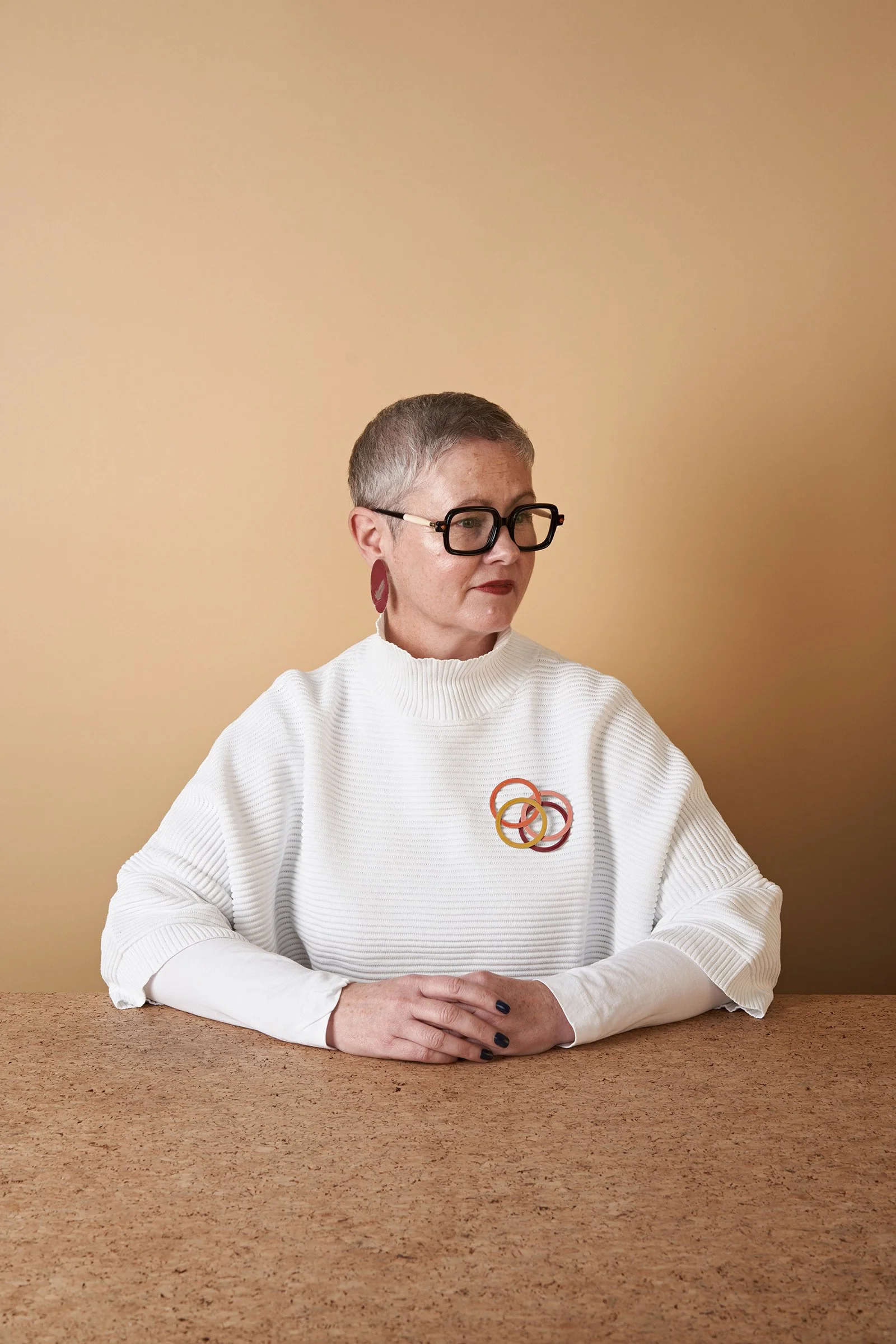 A woman with short gray hair and glasses sitting at a wooden table against beige background. She is wearing a white ribbed sweater with colorful loop pins on it and large earrings.