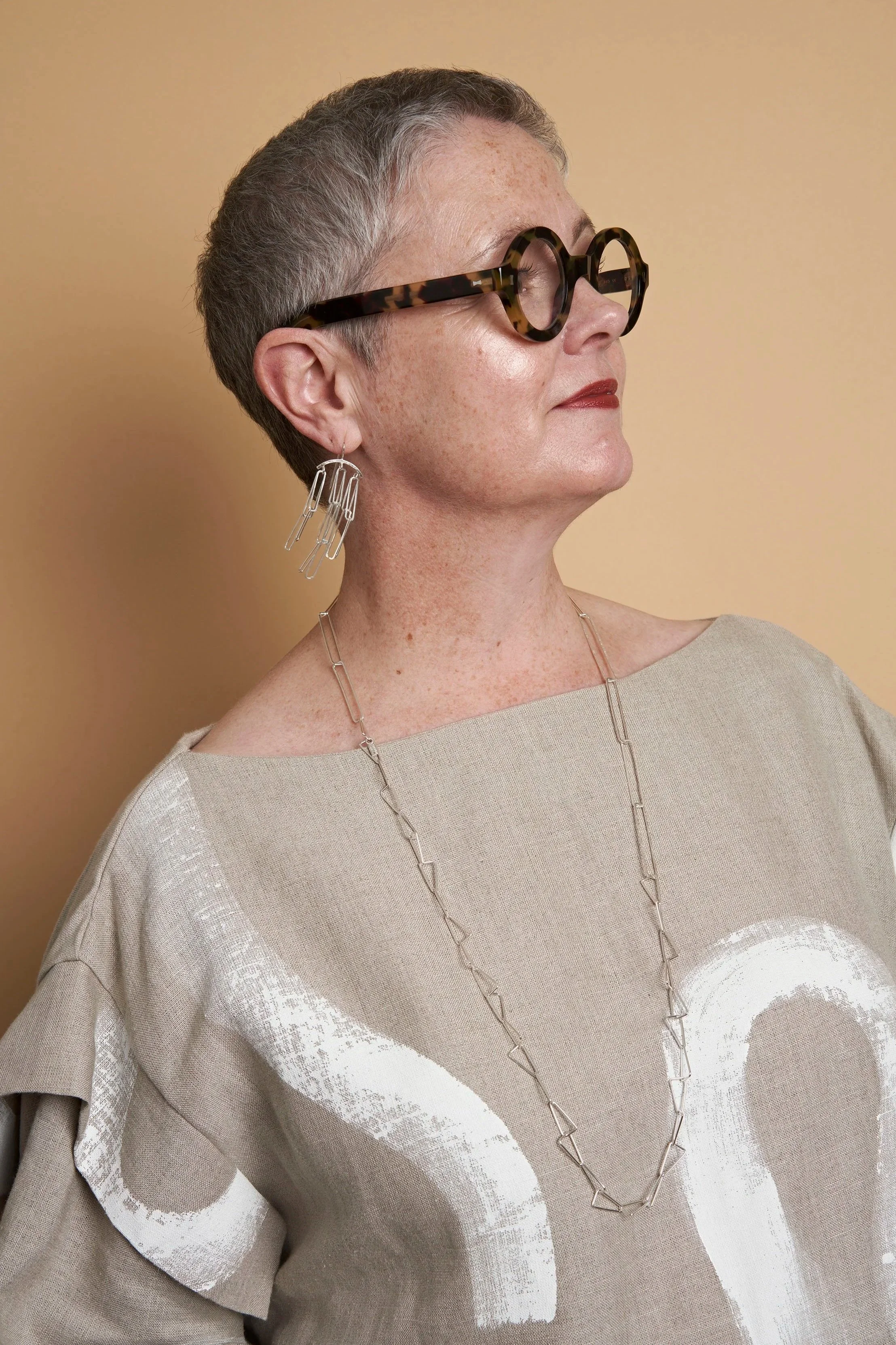 A woman with short gray hair wearing tinted round glasses, silver geometric earrings, a beige top with white abstract print, and a long silver necklace, standing against a plain beige background.