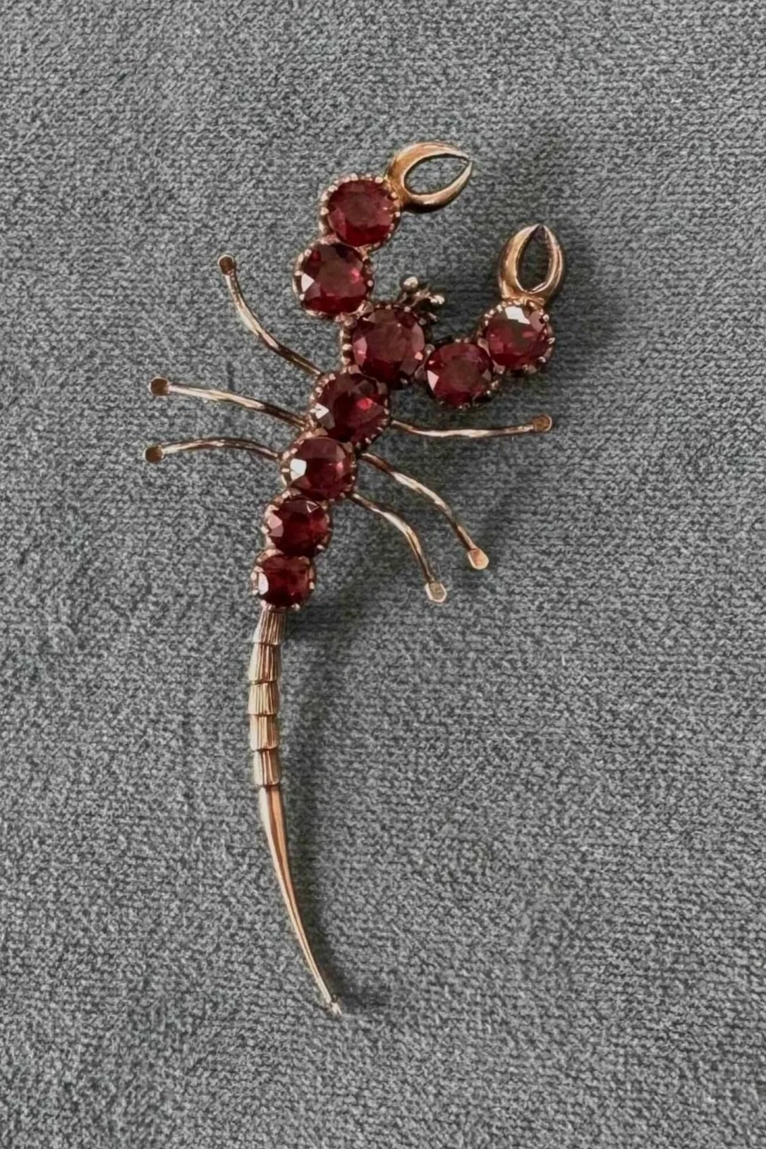 Gold-colored bug-shaped brooch decorated with red gemstones on a gray fabric background.