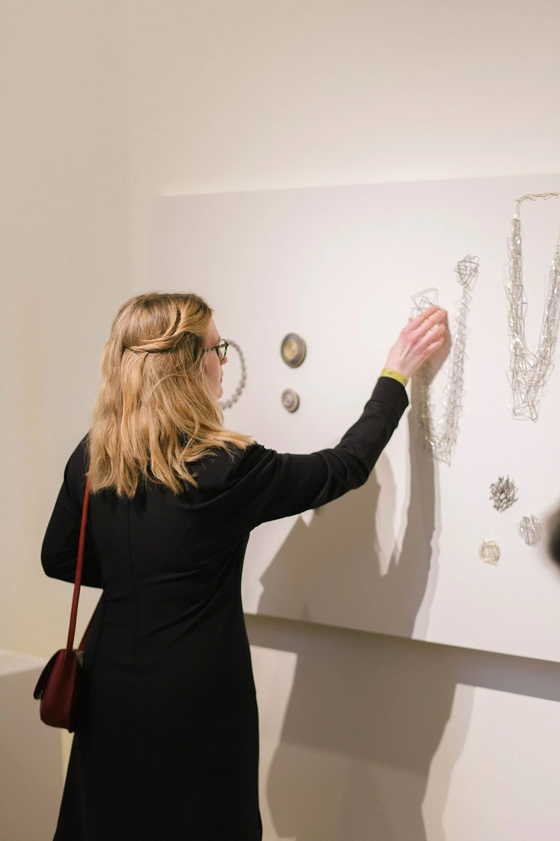 Woman with blonde hair and glasses wearing a black coat viewing metallic wire sculpture art on a gallery wall.