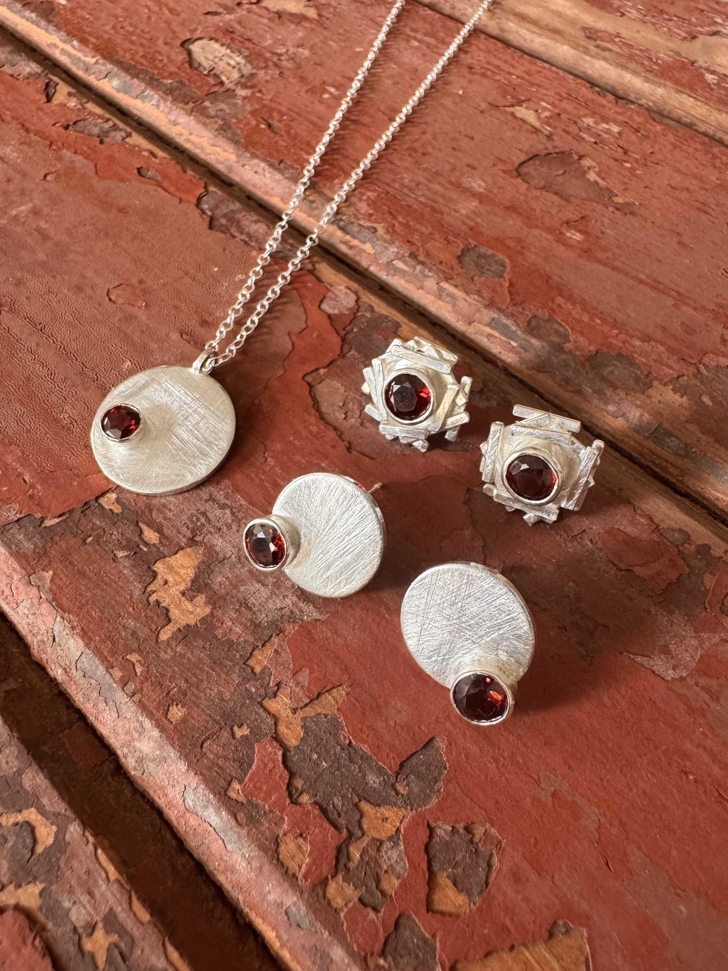 Silver jewelry set with red gemstones, including a necklace, earrings, and a ring, displayed on a rustic red wooden surface.
