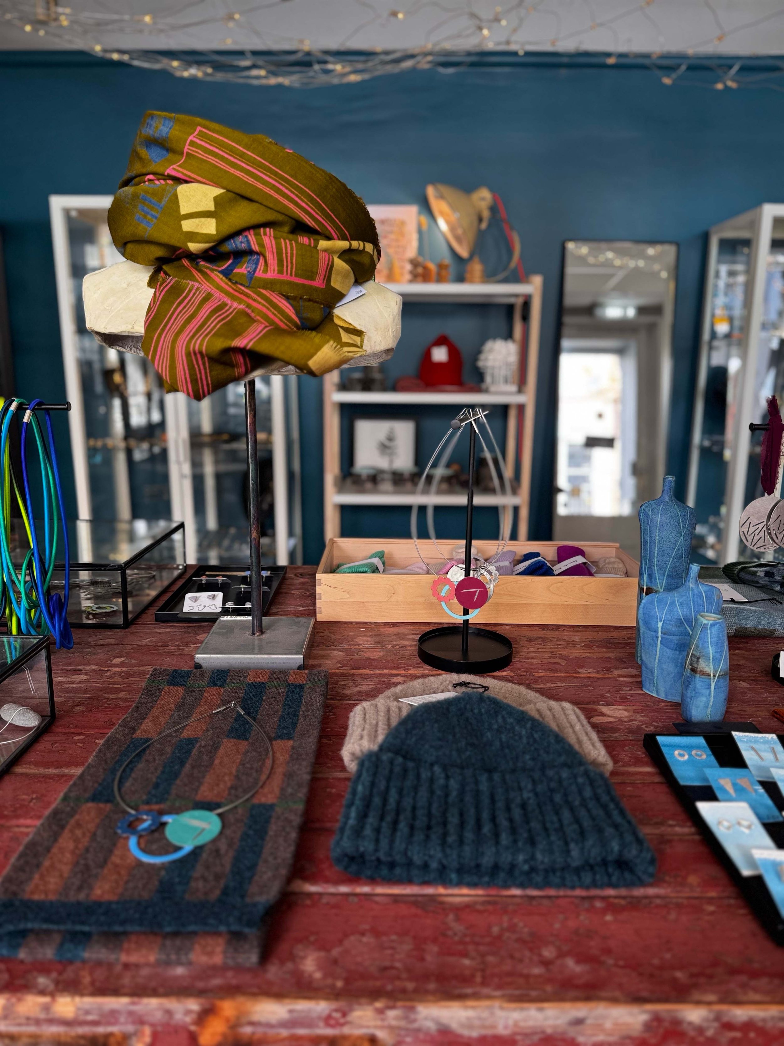 Various textile samples and jewelry displays on a wooden table in a boutique store, including scarves, fabric swatches, and jewelry rings with a blue wall and shelves in the background.