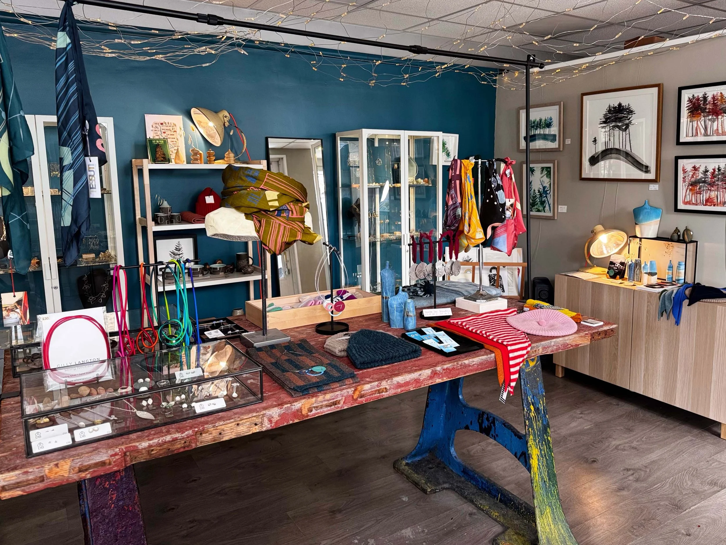 Interior of a retail store with shelves displaying jewelry, colorful scarves hanging on a clothing rack, and framed artwork on the walls.