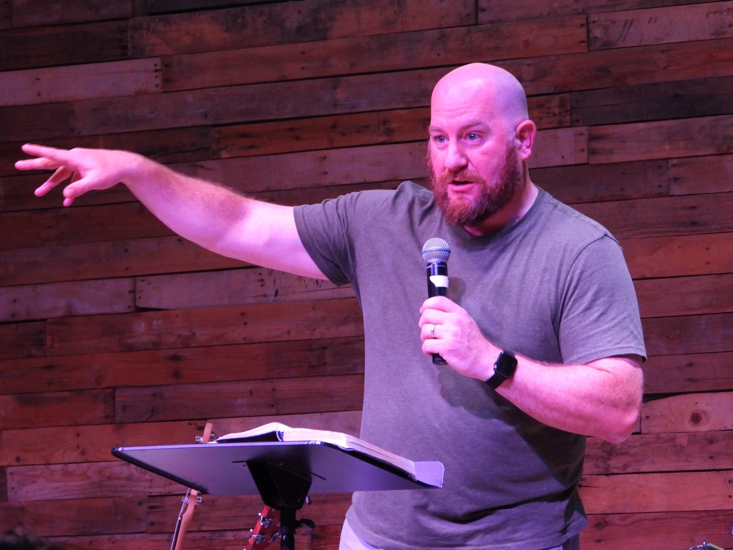 A man with a bald head and beard, wearing a gray t-shirt, holding a microphone and speaking, with a music stand and open notebook in front of him, against a wooden wall background.