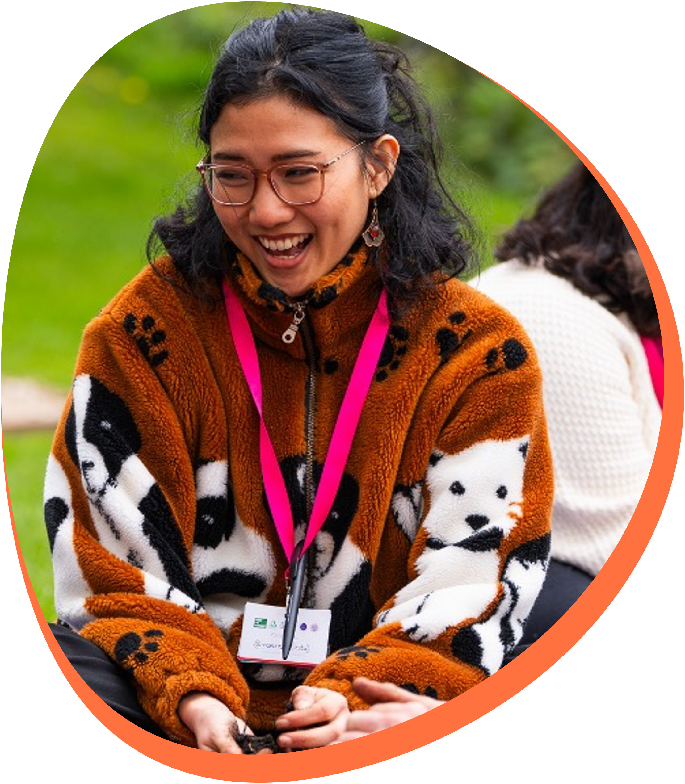 A young woman with glasses laughing, wearing a patterned fleece jacket, sitting outdoors with blurred greenery in the background. She has a lanyard with a name tag around her neck.