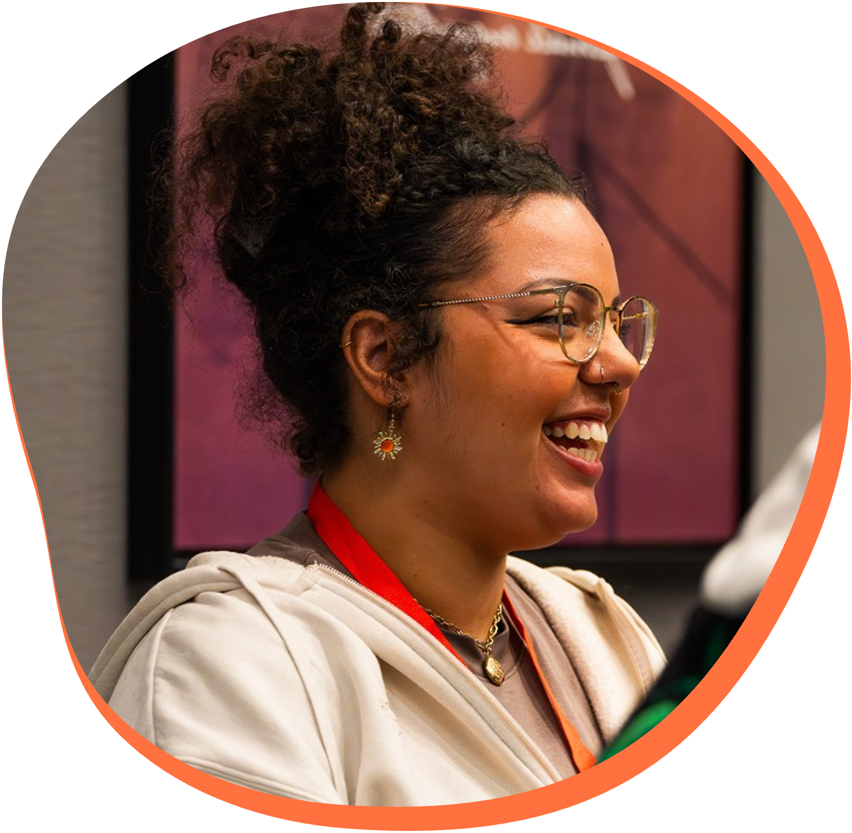 A woman with glasses and curly hair smiling, wearing a beige hoodie and jewelry, with a pinkish background.