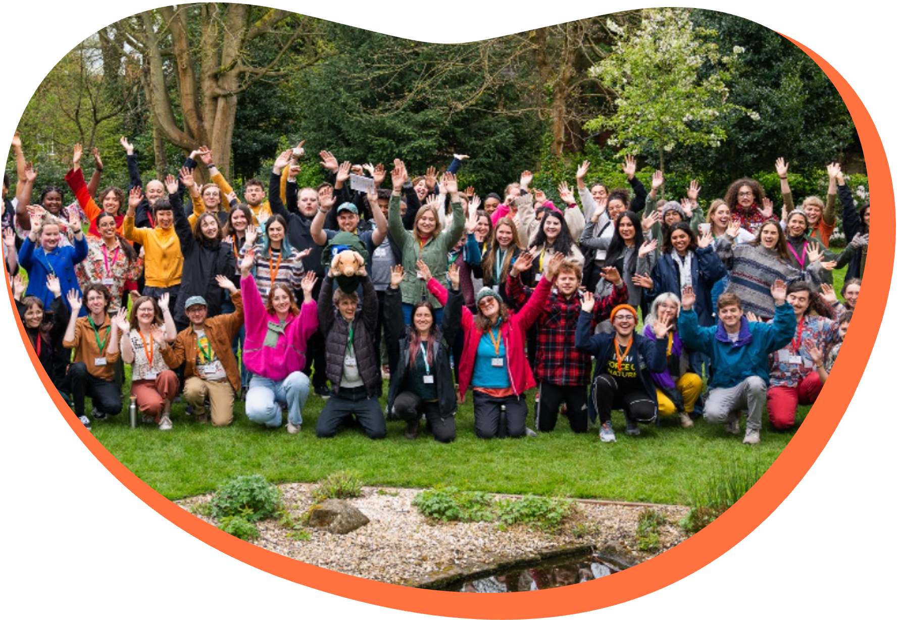 A large group of smiling people posing outdoors in a park with trees and grass, raising their hands in celebration.