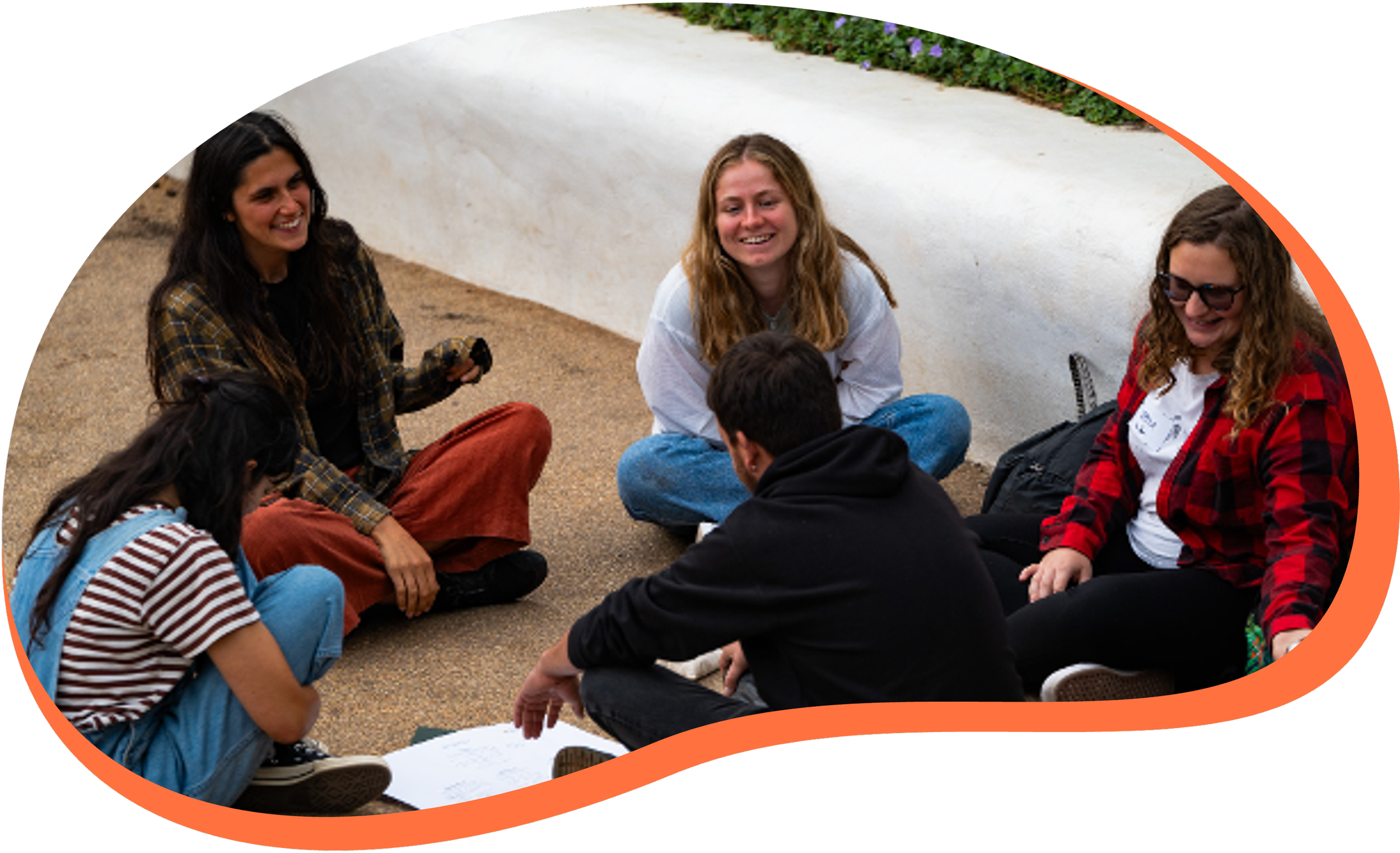 Group of young people sitting on the ground in a circle, smiling and engaging in conversation outside, near a white wall with green plants.