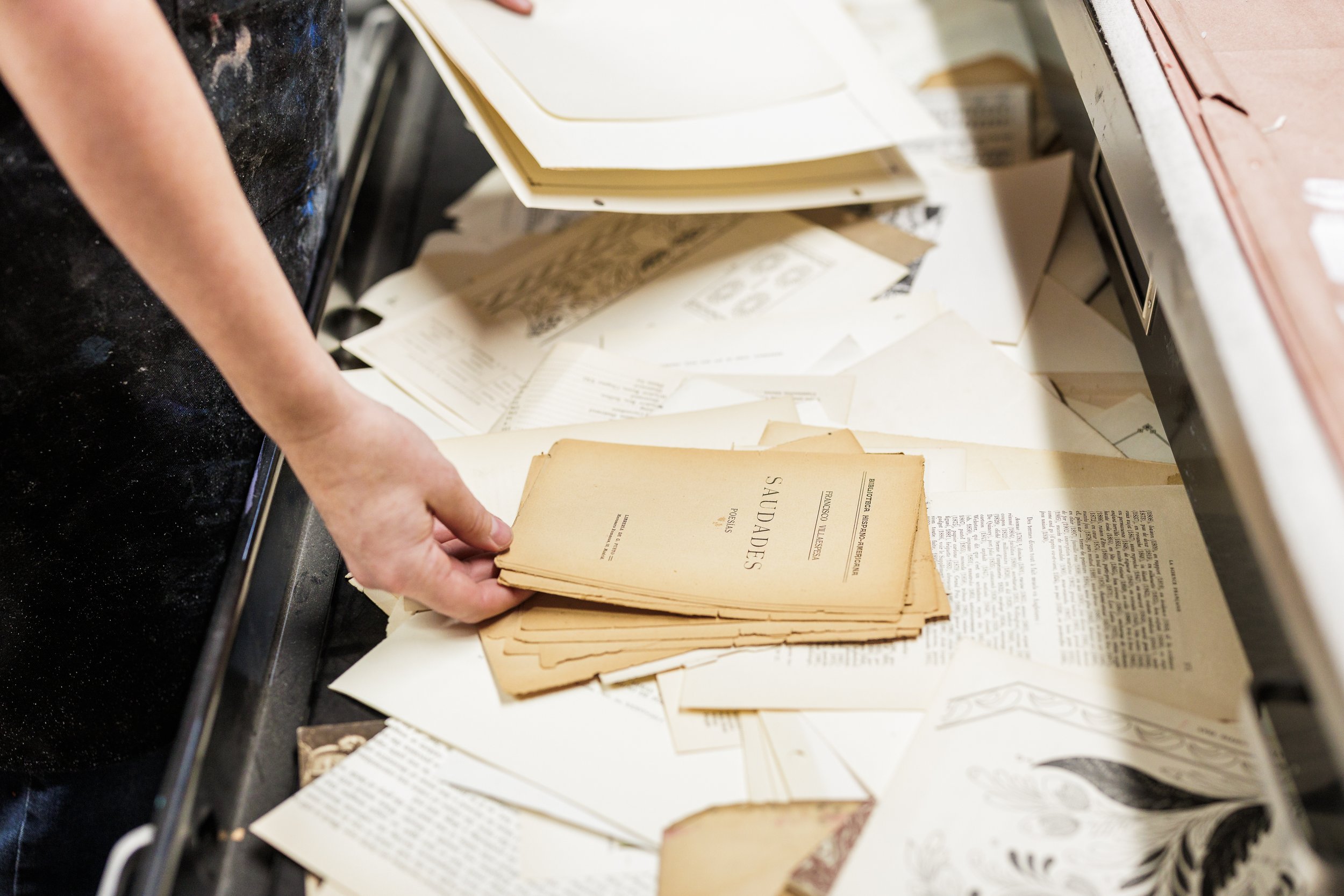 Person organizing vintage papers, including a document titled 'Saudades,' on a cluttered table.