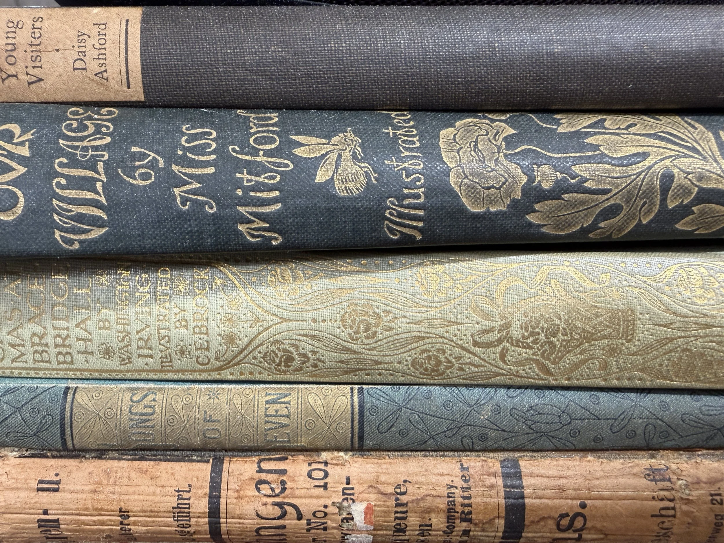 Close-up of several vintage-style books stacked horizontally, showing ornate gold and engraved designs on the covers, with visible text and detailed patterns.
