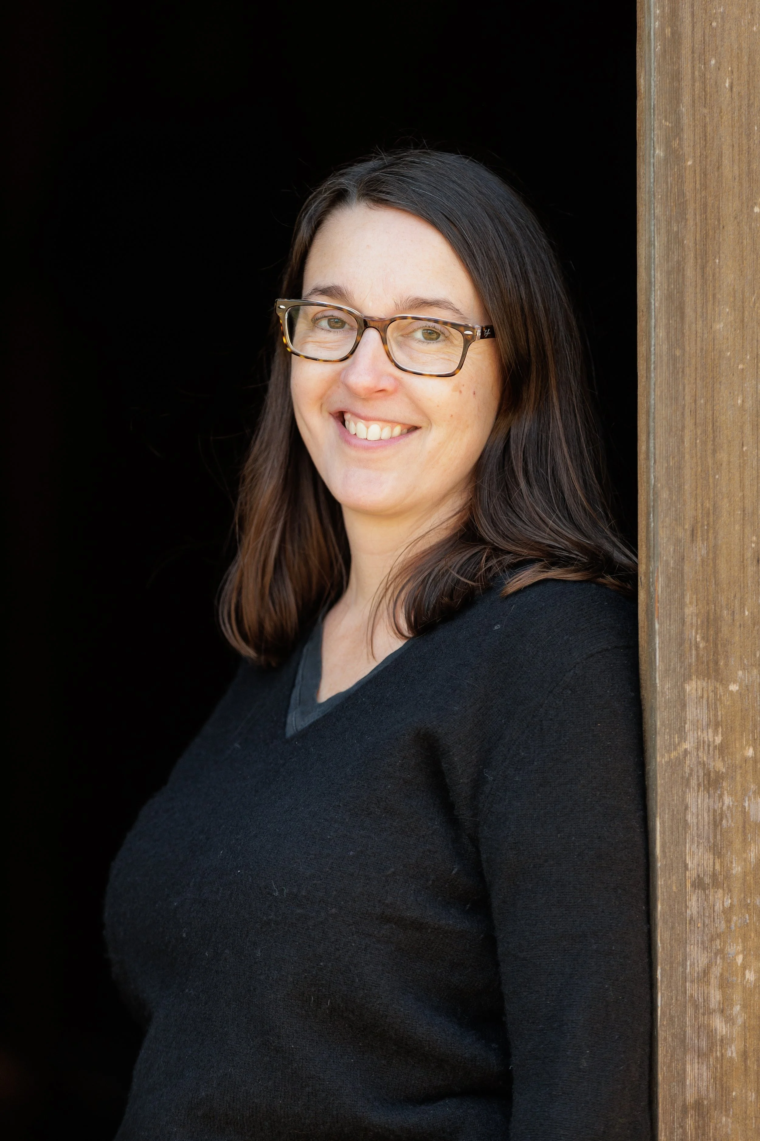 Woman with long brown hair and glasses smiling while leaning against a wooden surface, wearing a black top.