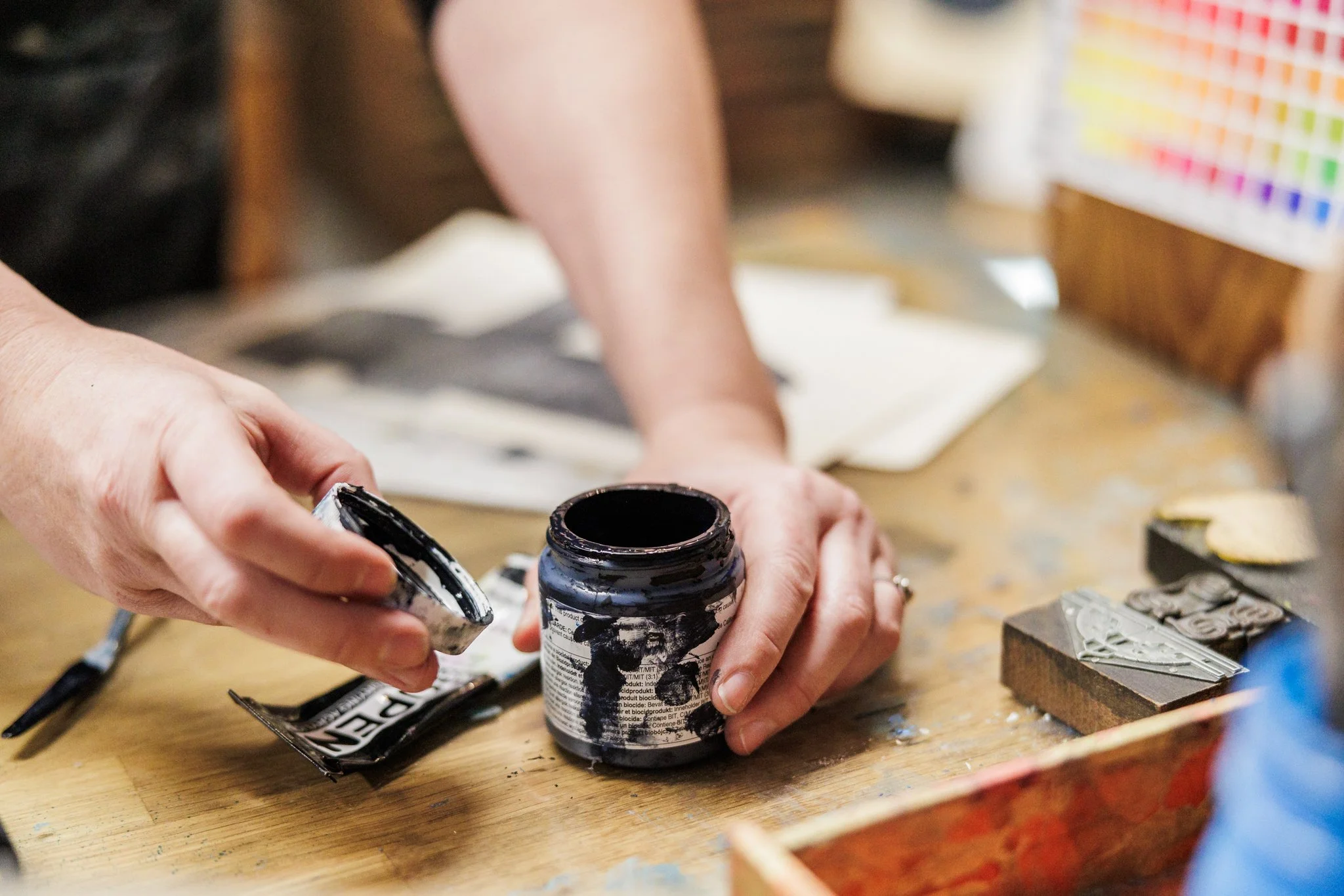 Hands opening a black ink or paint jar on a wooden table, surrounded by art supplies and papers.