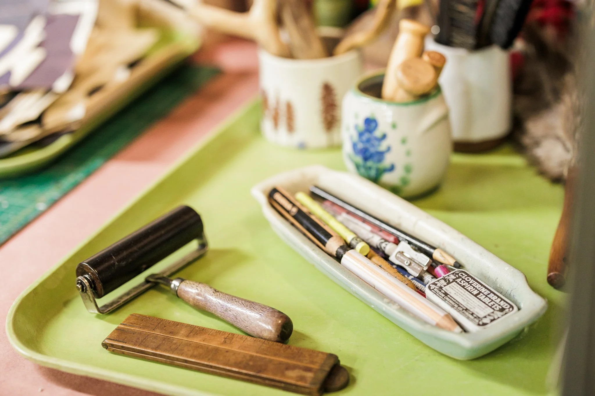 A green tray holding a lint roller, a wooden ruler, a ceramic dish with various colored pencils and a pen, and nearby ceramic containers with brushes and other tools.
