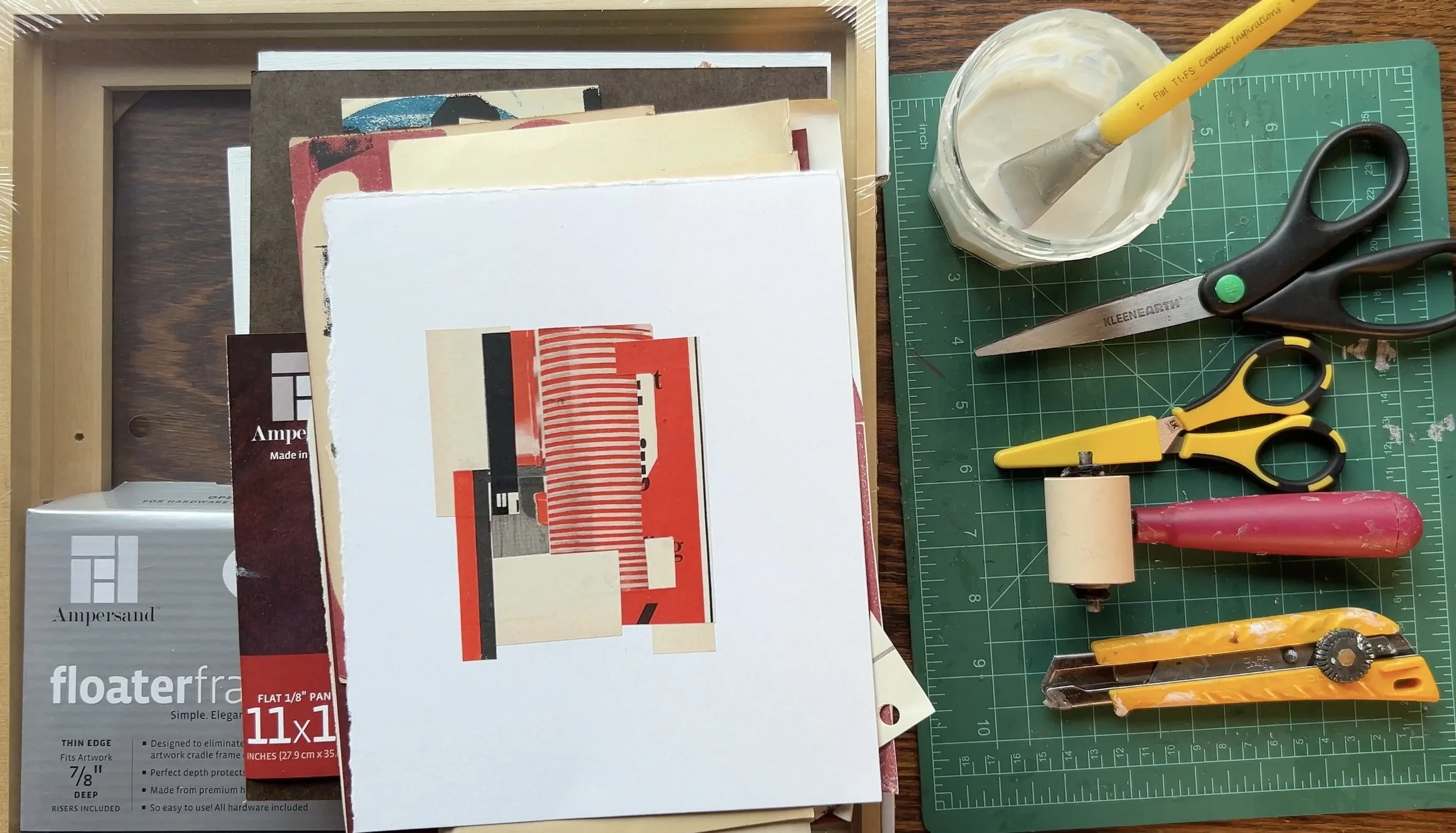 Art supplies on a table including a box of floater frames, mixed media papers, a cutting mat with scissors, a paintbrush in a jar, a roller, and a utility knife.