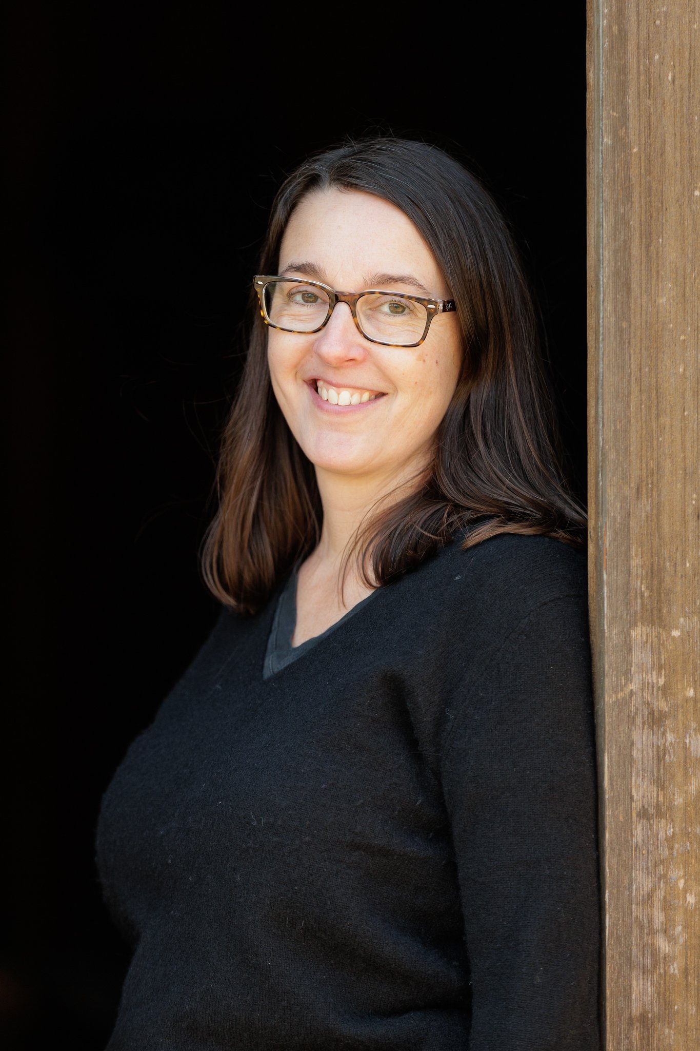 A woman with shoulder-length brown hair and glasses smiling while leaning against a wooden post, wearing a black sweater.