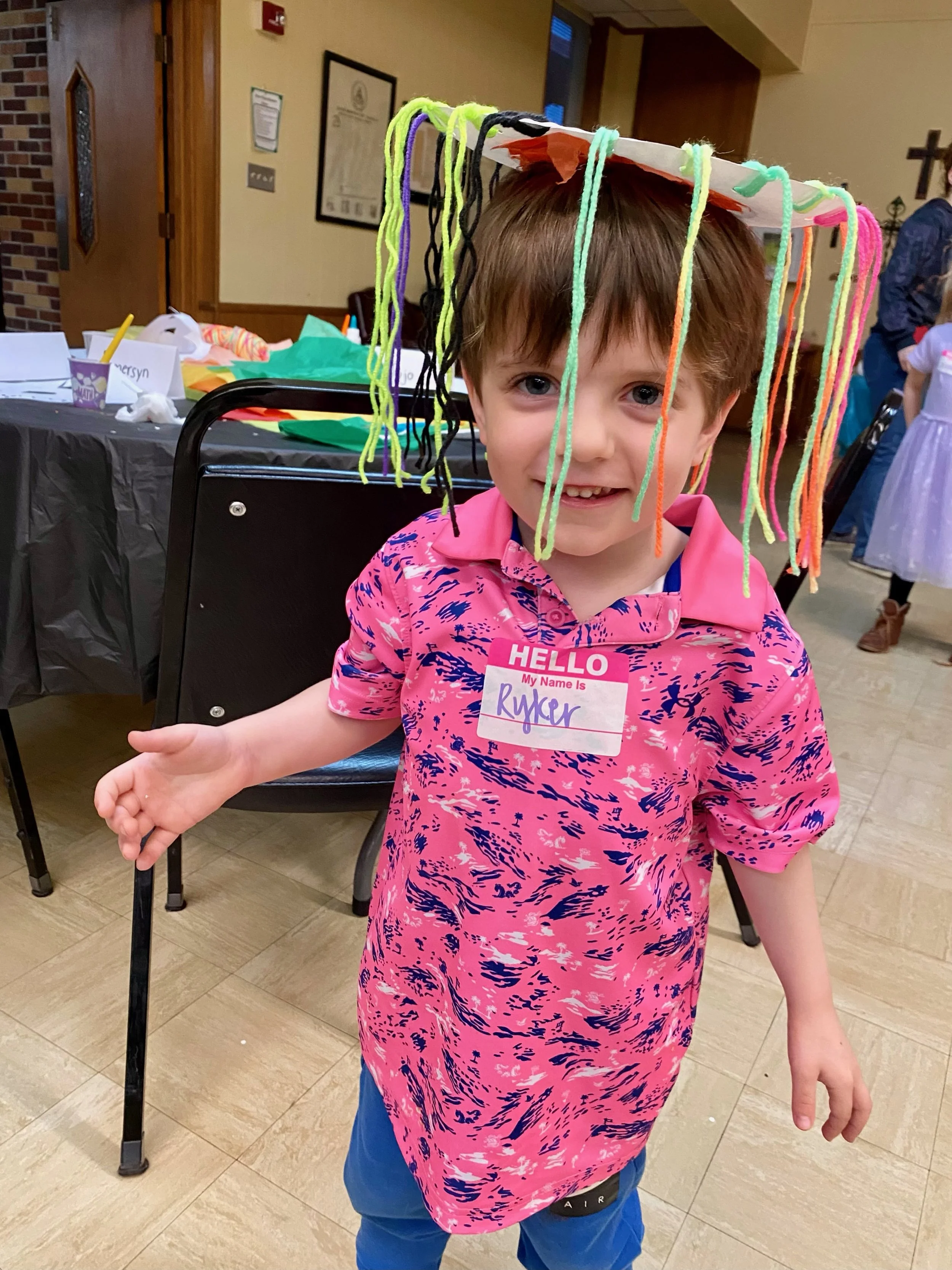 boy in a pink shirt has a paper plate with strings on his head