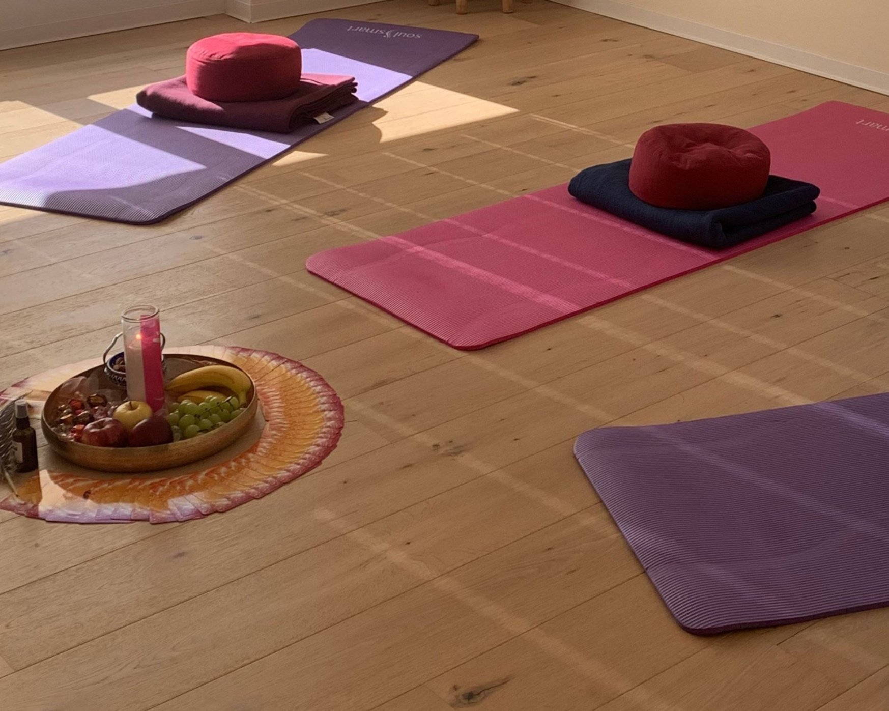 Yoga-Matten in verschiedenen Farben mit Polster, Handtüchern und einem Korb mit Früchten und Kerze auf einem Holzfußboden in einem Raum.
