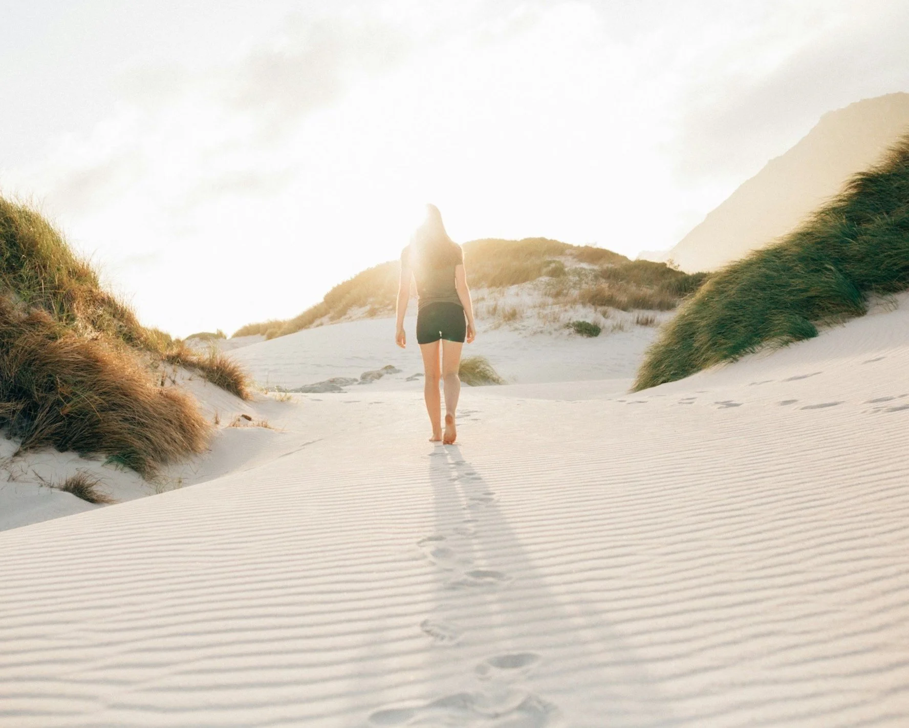 Eine junge Frau wandert barfuß auf einem Sandweg durch Dünen bei Sonnenuntergang, umgeben von Gräsern und Bergen im Hintergrund.