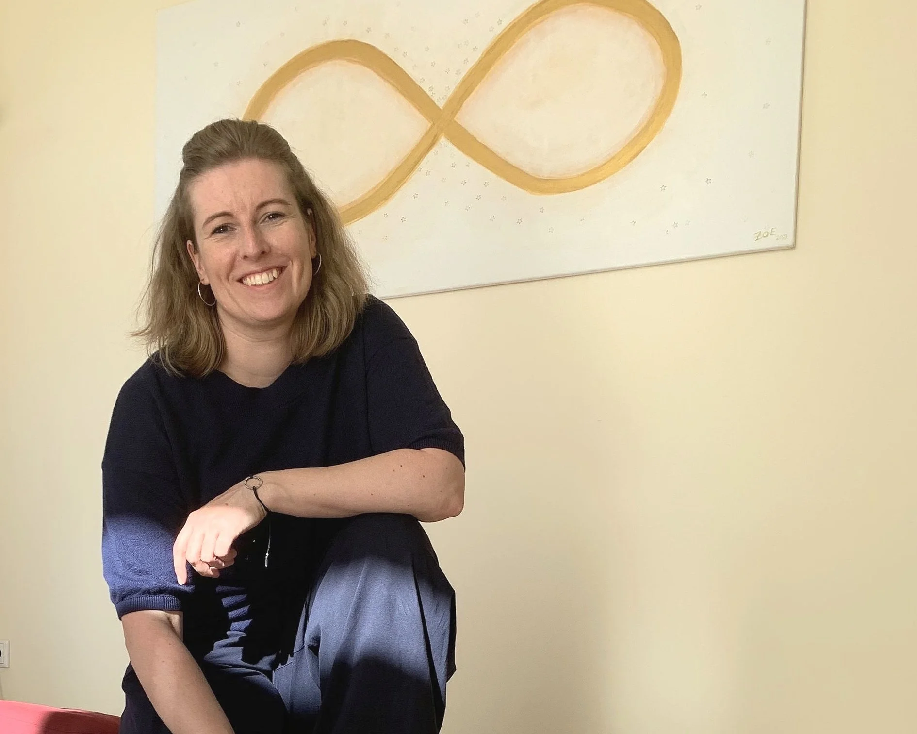 Freudige Frau mit braunen, gewellten Haaren, schwarze Bluse und dunkle Hose, sitzend auf einem roten Sofa vor einem weißen Wand mit abstrakter goldfarbener unendlicher Schleife Kunst.