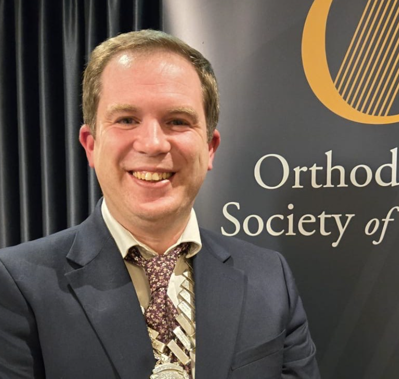 A man in a suit smiling at a conference with a banner behind him reading 'Orthodox Society of'.
