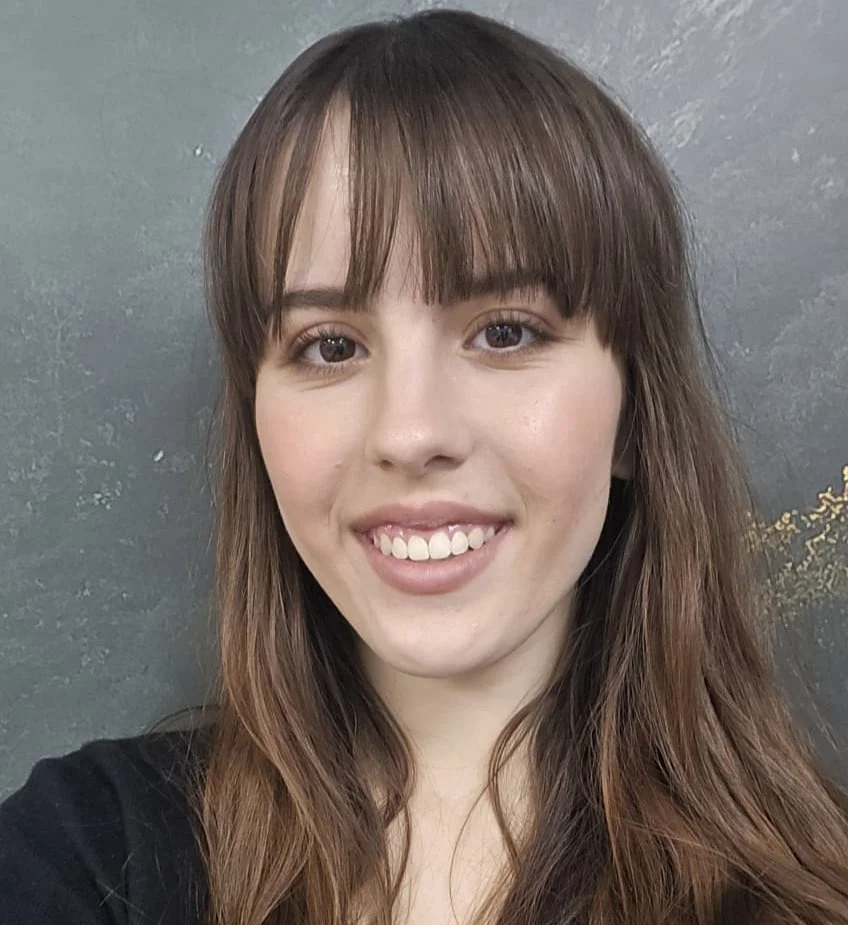 A young woman with long brown hair, bangs, and brown eyes smiling at the camera, wearing a black top, against a textured gray background.