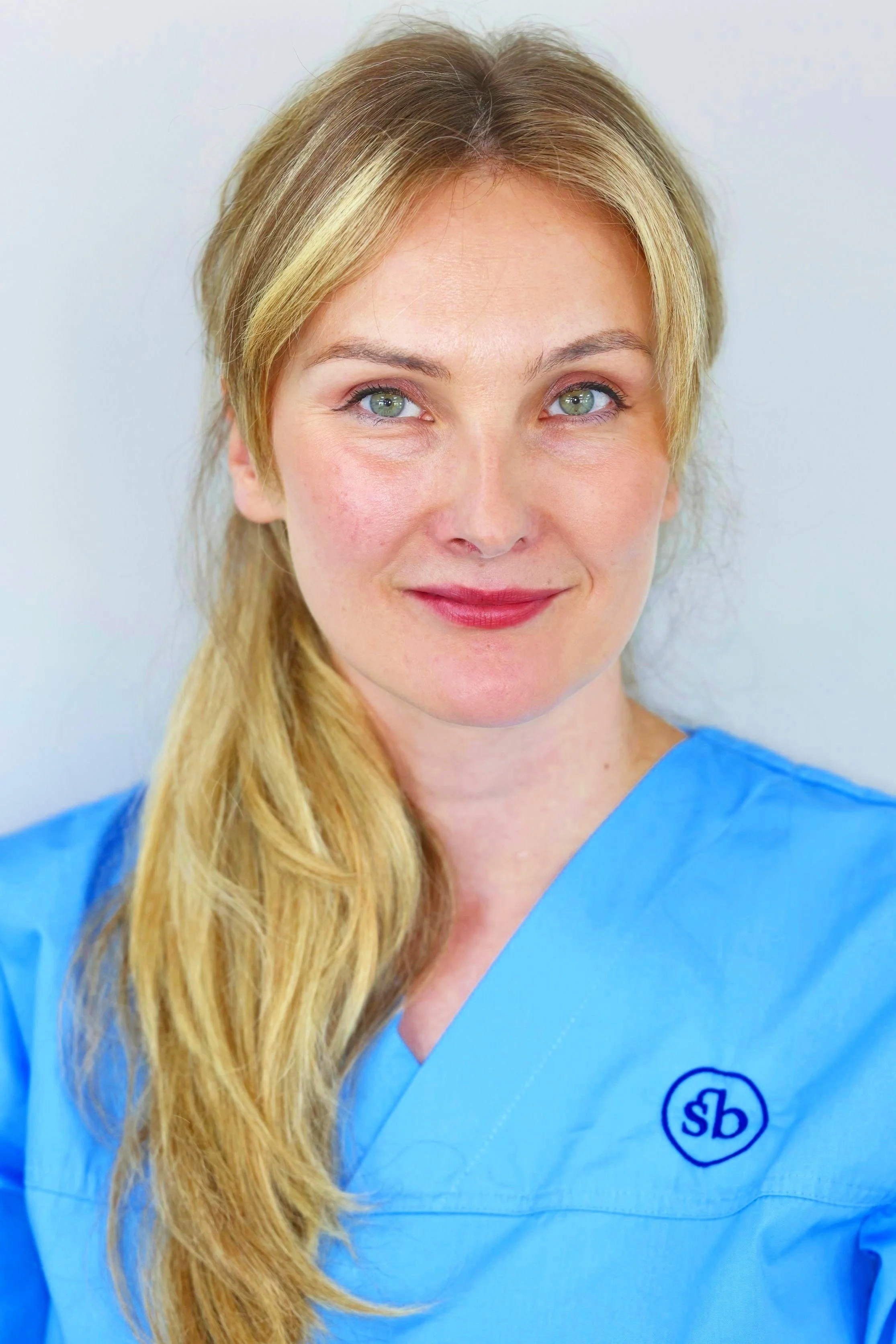 A woman with blonde hair and fair skin wearing blue medical scrubs, smiling, against a plain light-colored background.