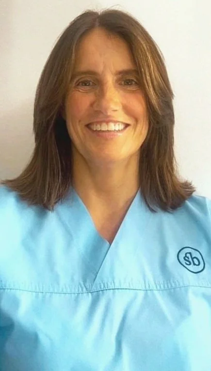 A woman with shoulder-length brown hair and a bright smile wearing light blue medical scrubs.