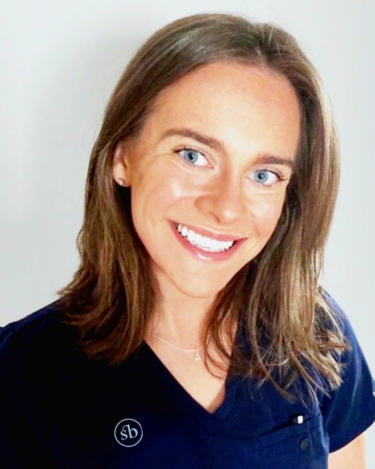 A woman with shoulder-length brown hair and blue eyes smiling, wearing a dark blue top with a small logo and a necklace with a letter A, against a plain white background.