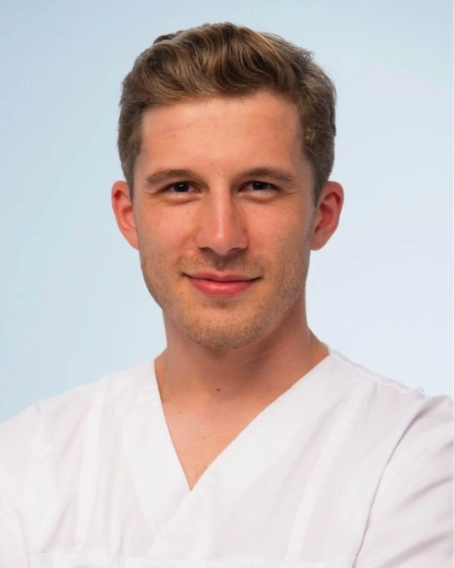 A young man with short, light brown hair, wearing a white medical coat, poses for a portrait against a light blue background.