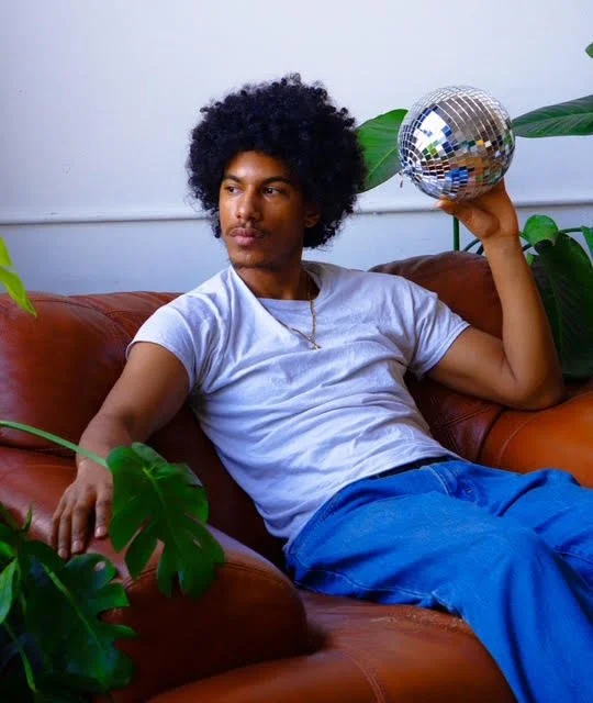 Isaiah Keaton sitting on a brown leather couch, holding a disco ball in his right hand, surrounded by green plants.
