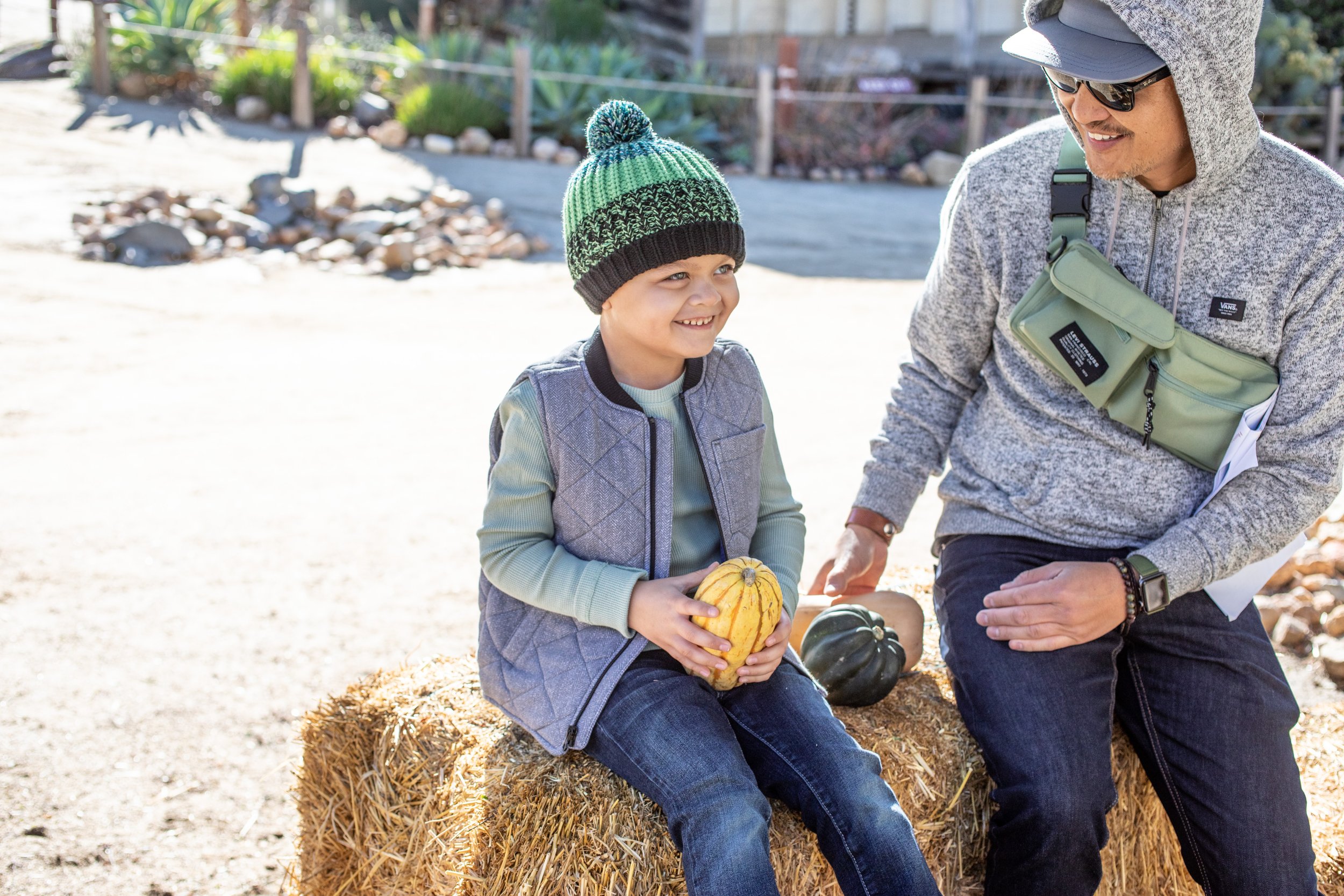 A man is sitting next to a boy on a hay stack at Leo Carrillo in Carlsbad California. The boy is wearing a green multi color beanie with a pom from San Diego Hat Company. 