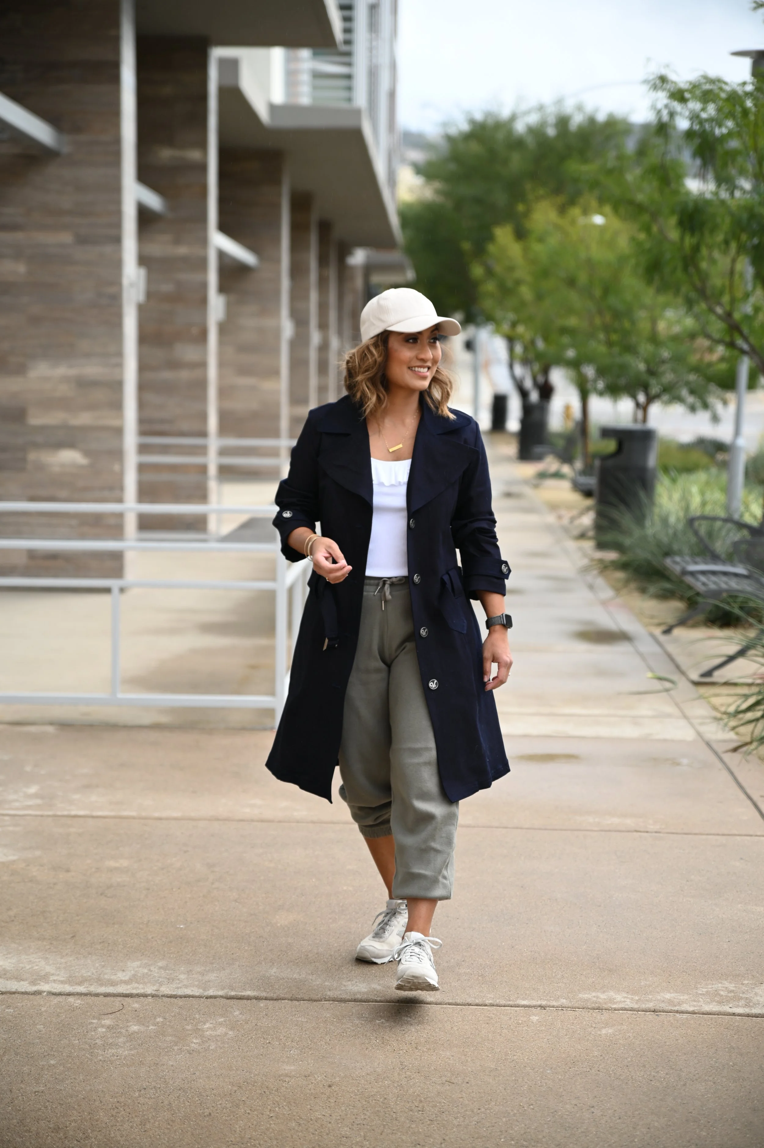 A happy woman walking down the street wearing a ball cap, white top, green sweatpants, denim trenchcoat, and New Balance sneakers.